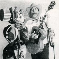 Ray Owen surrounded by numerous hats from various historical periods while playing guitar, harmonica and banjo